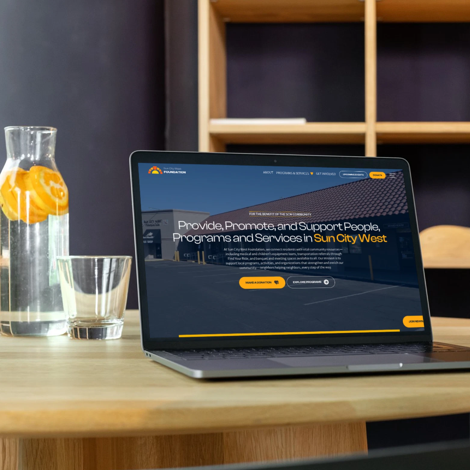 A laptop showing the Sun City West Foundation website beside a glass pitcher of orange-infused water on a wooden desk.
