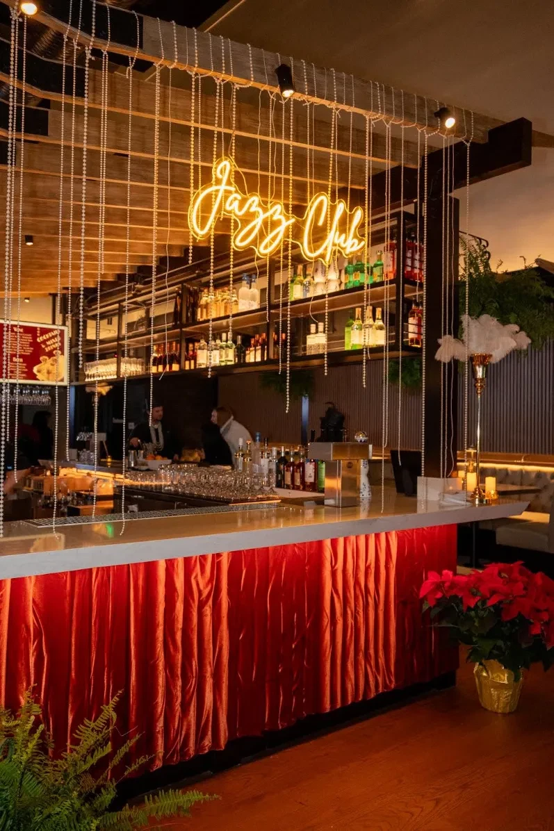 Speakeasy-style jazz club bar with a glowing yellow "Jazz Club" neon sign, cascading strands of beaded lights, a red velvet bar front, stocked liquor shelves, and red poinsettias beside ostrich-feather accents.