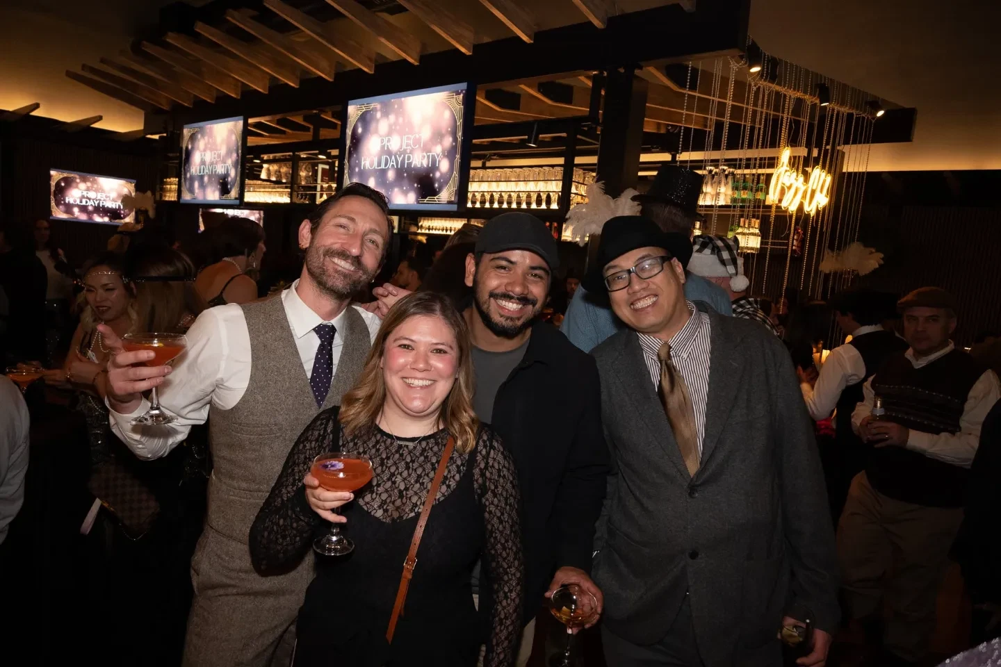 Four smiling guests in Great Gatsby–themed attire holding cocktails at a private holiday party, with "Project Holiday Party" screens and a neon sign glowing behind them.