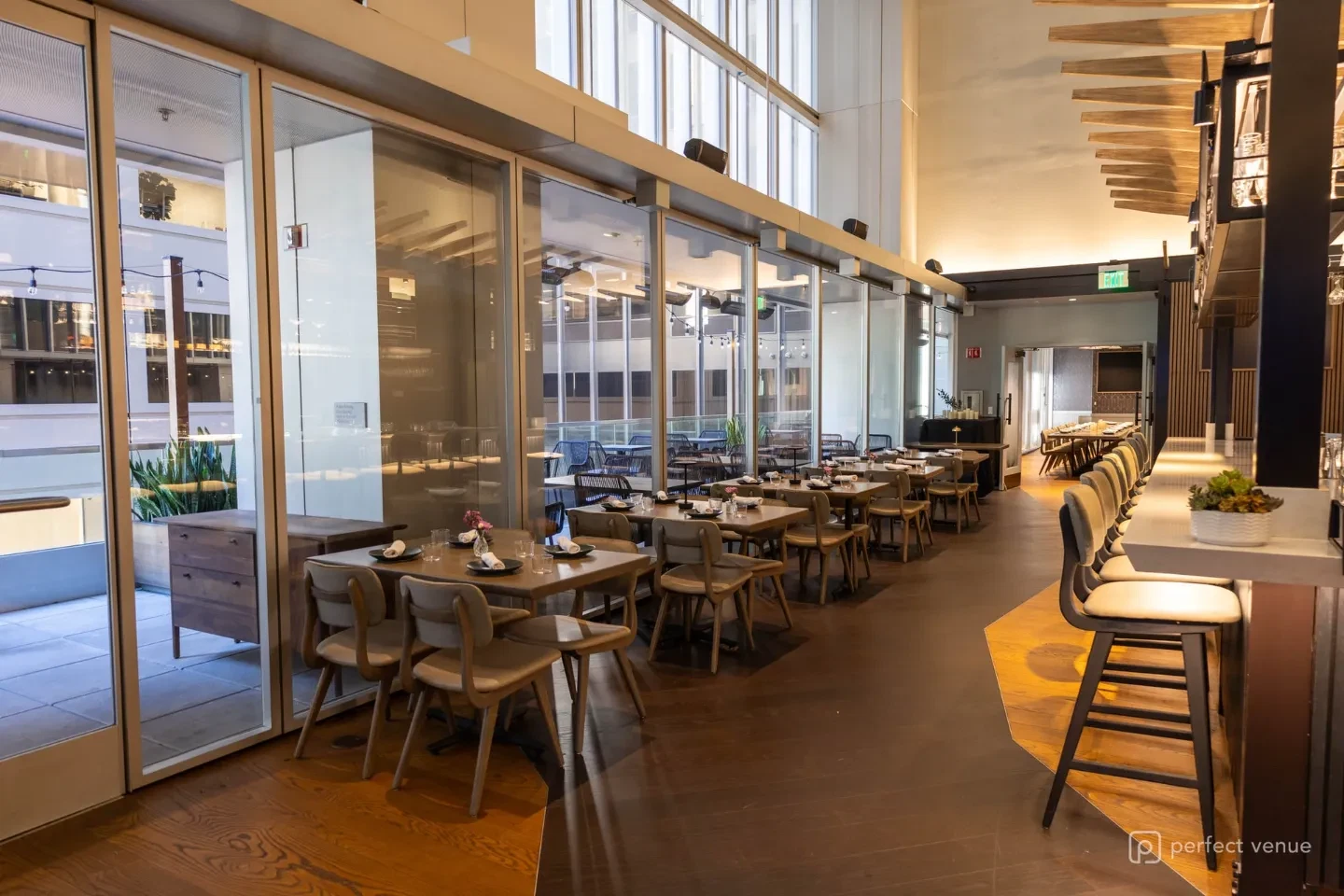 Upscale main dining room with floor-to-ceiling windows, wooden tables set with linen napkins, leather chairs, and a wood-floor pathway leading to the bar.
