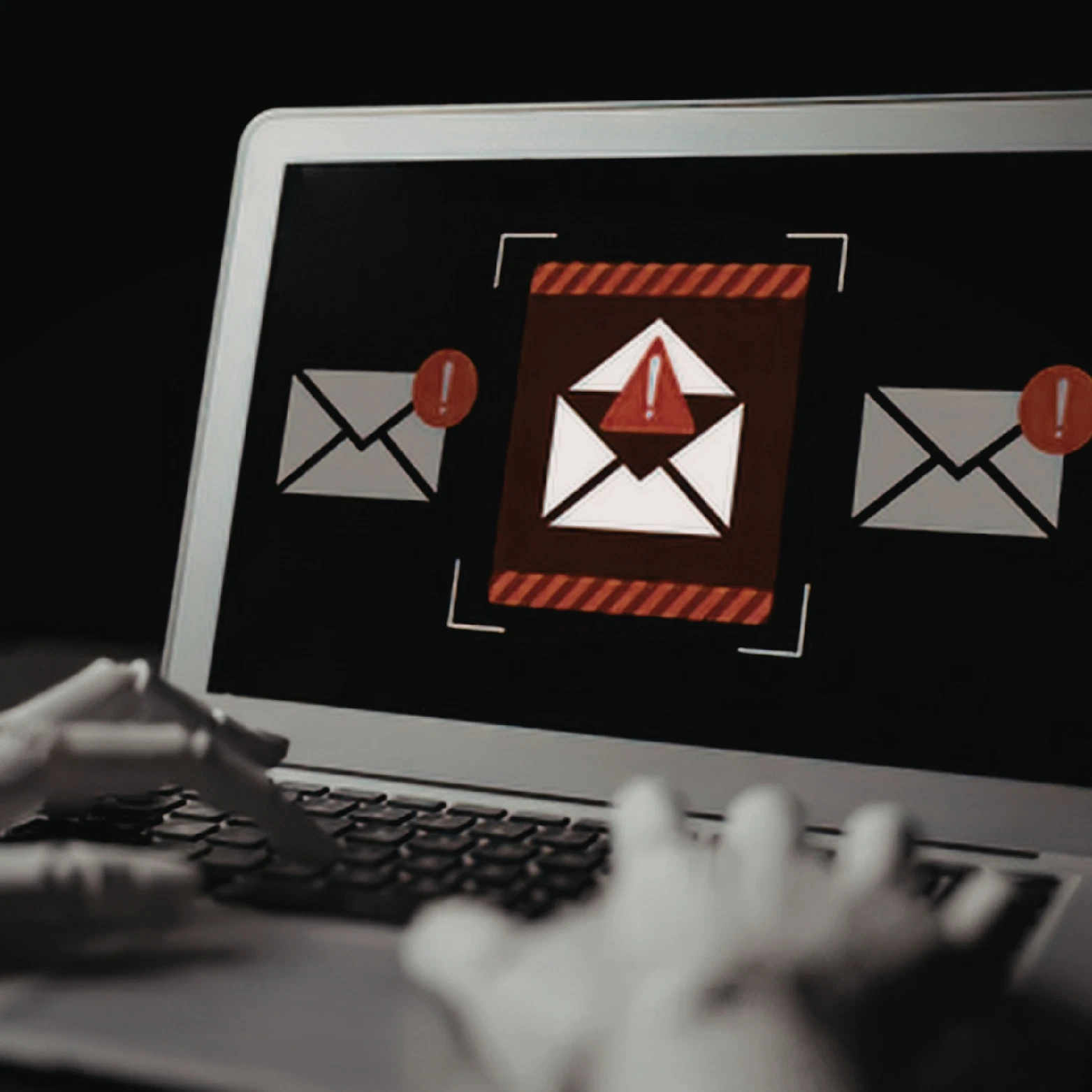 Hands typing on a laptop showing an alert symbol on an email icon, a reminder to watch for scams.