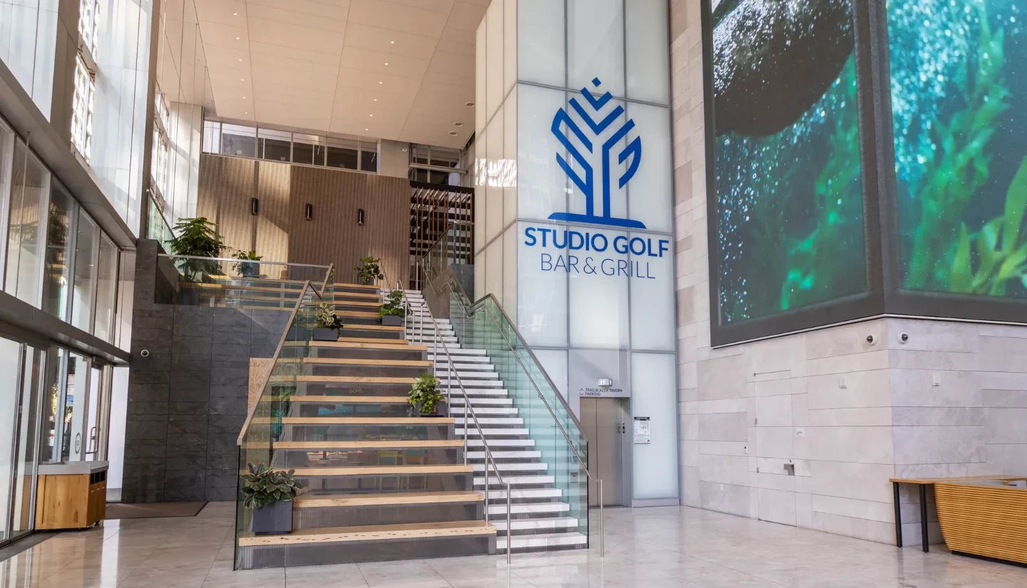Bright modern lobby entrance to Studio Golf Bar & Grill, featuring a wide glass-railed staircase lined with planters, marble walls, and a large blue logo sign next to a vibrant kelp-forest video display.