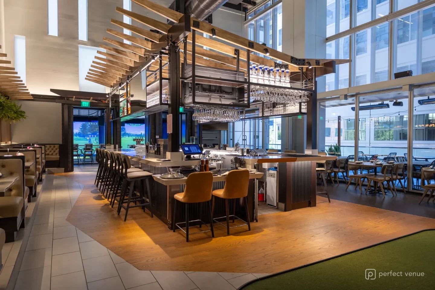 Modern restaurant bar with a suspended wood-slat canopy, hanging stemware racks, leather banquette booths, and a long row of upholstered bar stools.
