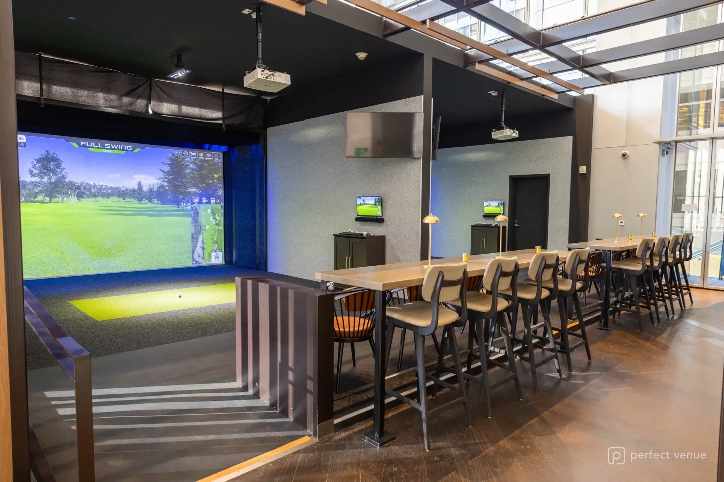 Indoor Full Swing golf simulator bay with a projected fairway, hitting mat, and a long communal high-top table lined with cushioned bar stools.