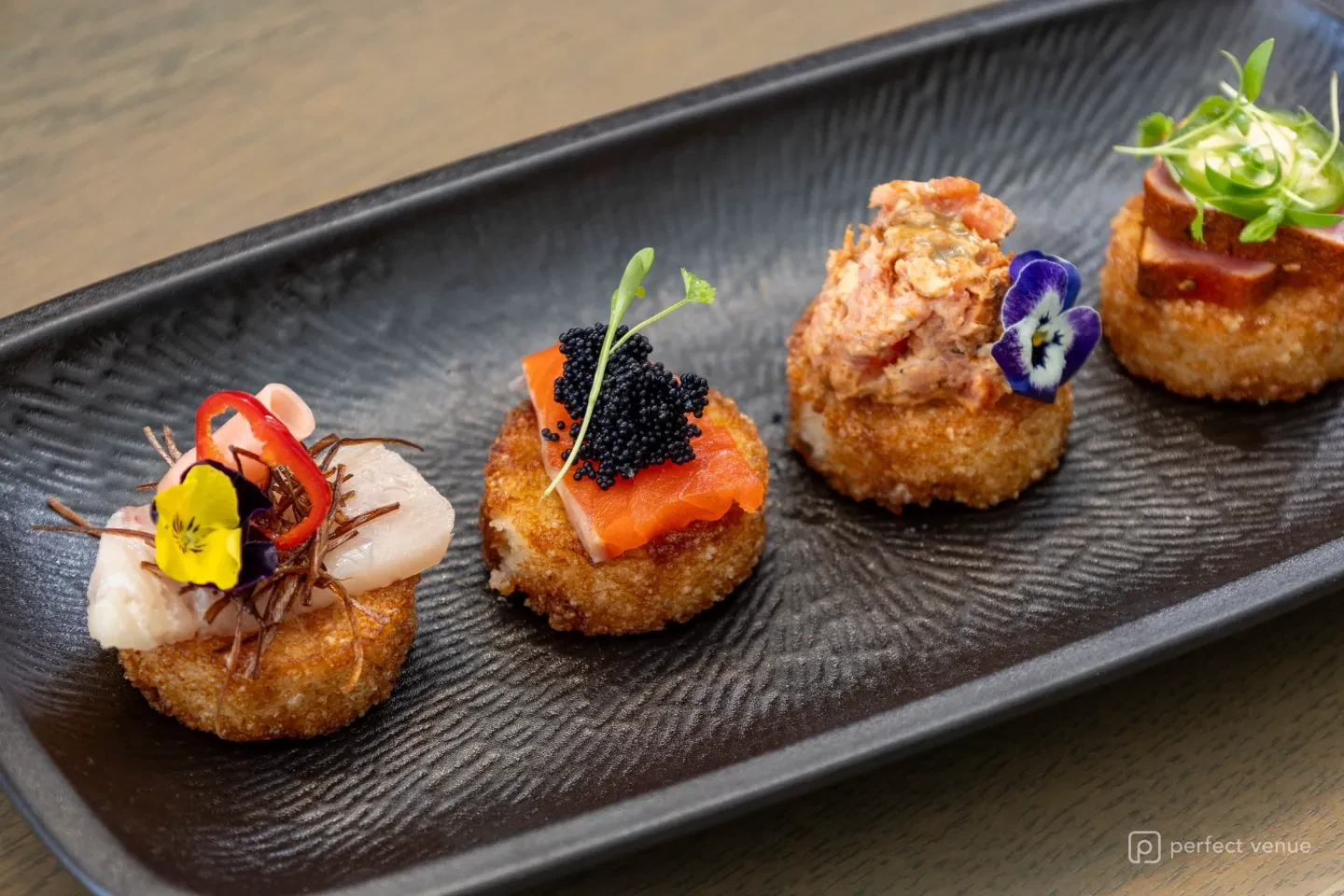 Four bite-sized gourmet canapés on a textured black ceramic platter, garnished with caviar, edible flowers, microgreens, and sliced peppers.