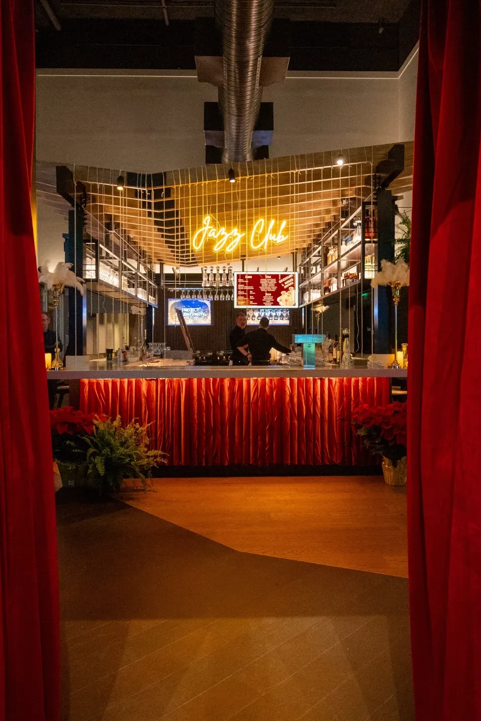 Entrance to an intimate jazz club framed by red velvet curtains, revealing a copper-fronted bar, a glowing yellow "Jazz Club" neon sign, and bartenders mixing drinks.