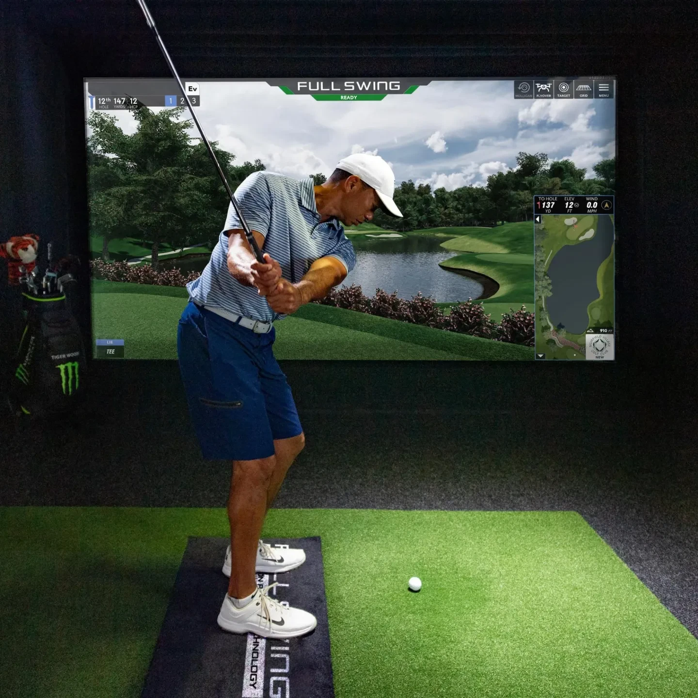 Golfer in a white cap, striped polo, and navy shorts mid-backswing on a Full Swing simulator bay, with a scenic virtual course and shot tracking data displayed on the screen.