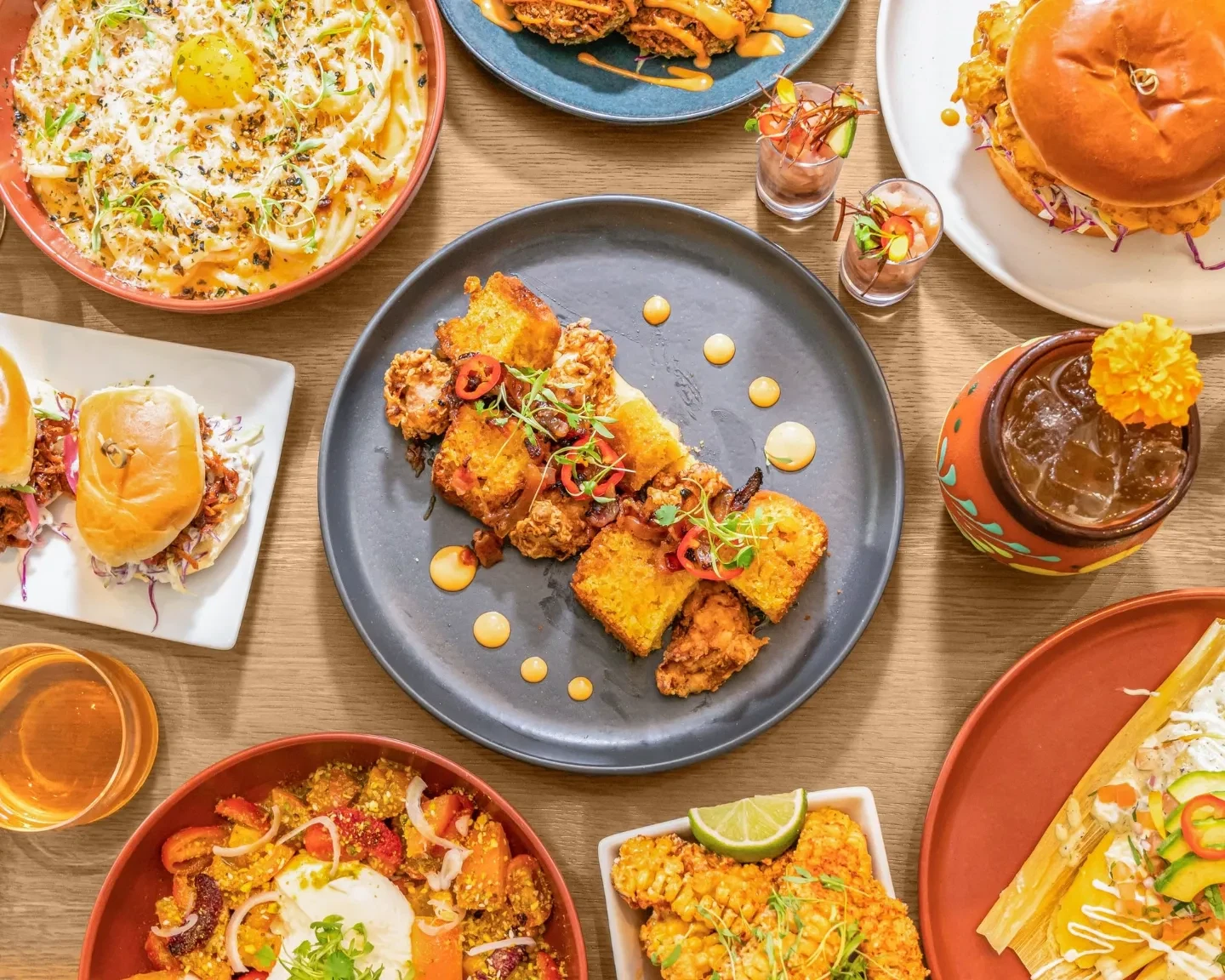 Overhead view of a Latin-inspired shareable spread featuring crispy fried chicken, sliders, loaded nachos, a smashed burger, ceviche tostadas, and craft cocktails on a wooden table.