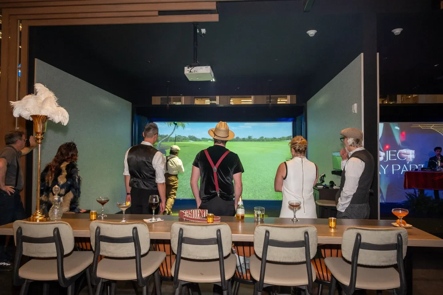 Guests in 1920s Gatsby-themed attire watching a swing on a large Full Swing golf simulator screen during a private event, with cocktails lined up on a high-top counter and ostrich-feather decor in the foreground.