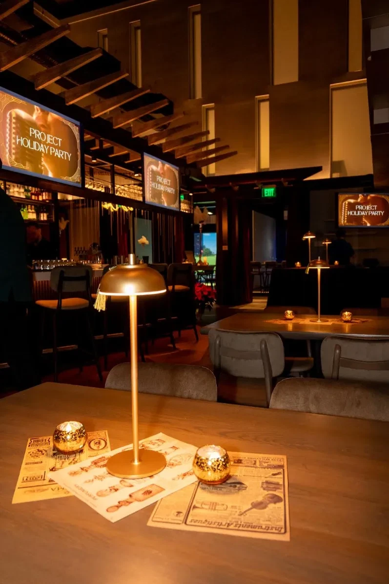 Intimate private event seating with a brass dome table lamp, gold disco-ball votives, and vintage-style newspaper place mats, set against glowing "Project Holiday Party" screens in the background.