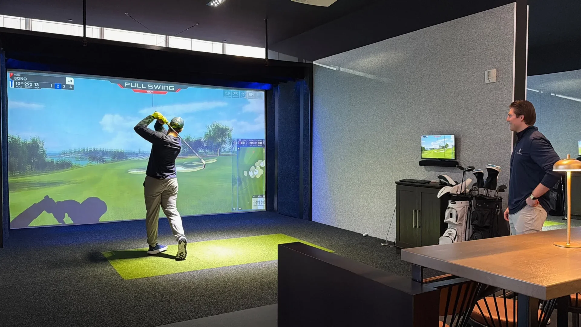 Golfer mid-follow-through on a Full Swing simulator bay, with a coastal virtual course displayed on the large screen showing a "10th hole, 292 yards" scoreboard, while a teammate in a navy pullover watches from beside a rack of golf bags and a high-top table.