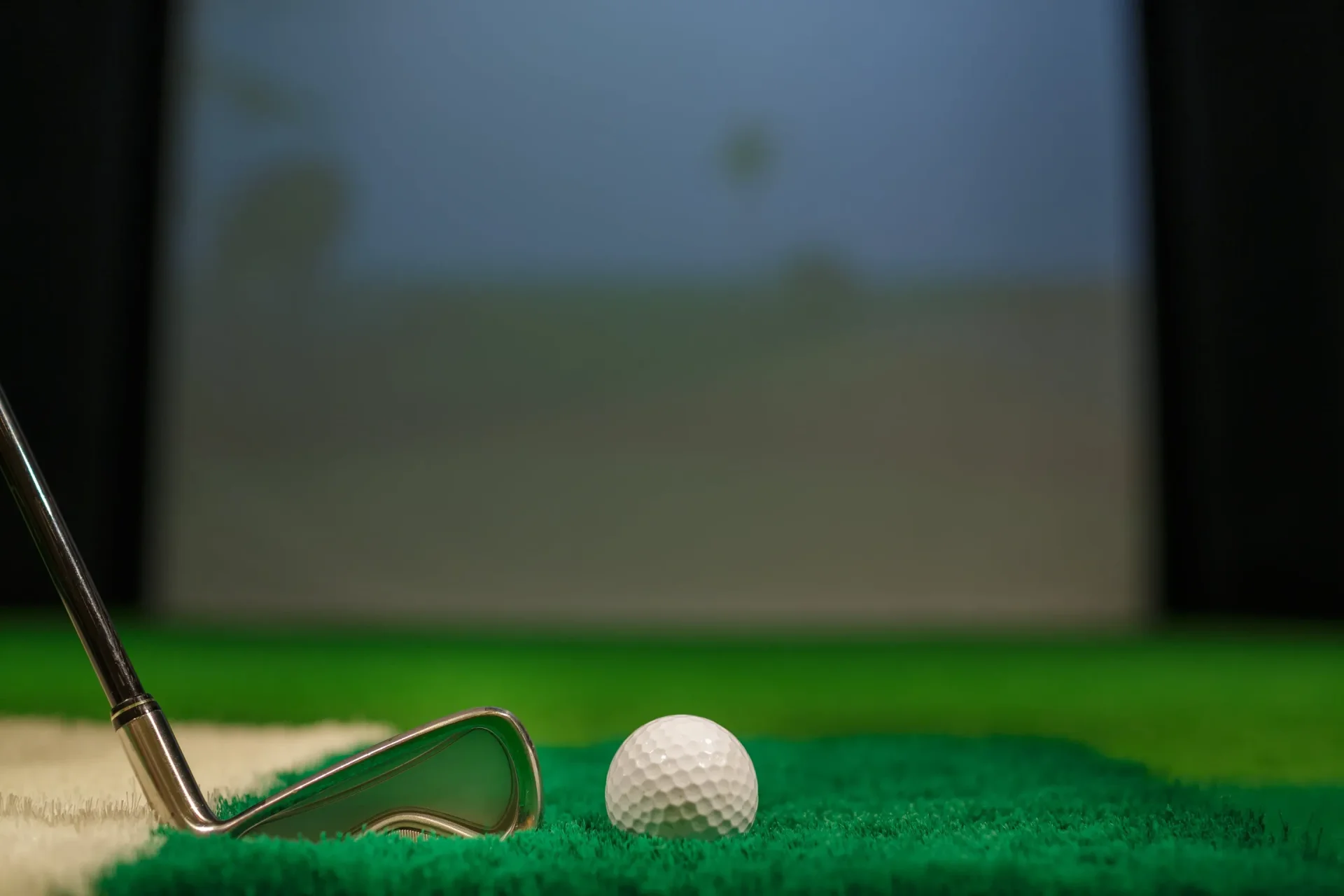 Close-up of a polished iron golf club positioned next to a white golf ball on a synthetic turf hitting mat in front of a softly lit simulator screen.